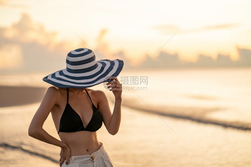 身穿比基尼和白色长裤的顶层比基尼女人在海滩上戴帽子背面日出或落美丽者乐趣海洋图片
