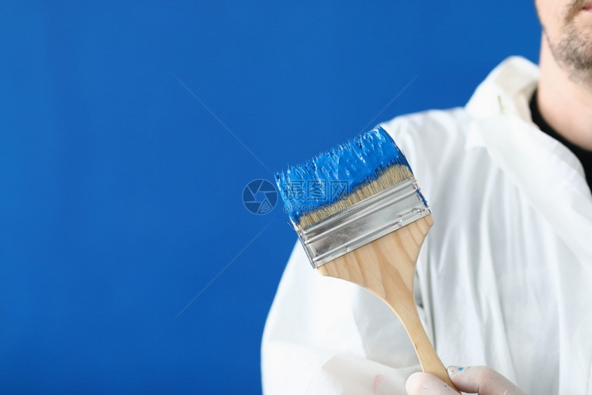 身穿白色防护服的男子手里拿着画笔蓝墙上涂有色油漆的木刷身穿白色防护服的男子手里拿着画笔男人套装阴凉处图片