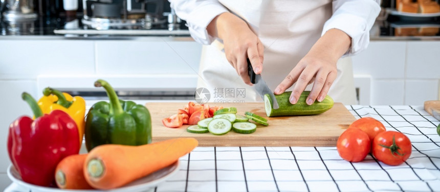 生的亚洲家庭主妇穿围裙用刀在切削纸板上黄瓜和番茄同时准备配料和烹饪供健康餐的午时在家厨房吃尽管沙拉图片