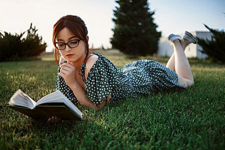 微笑铺设复古的身着老代旧礼服天主教女孩在日落时读了一本关于草坪的书图片
