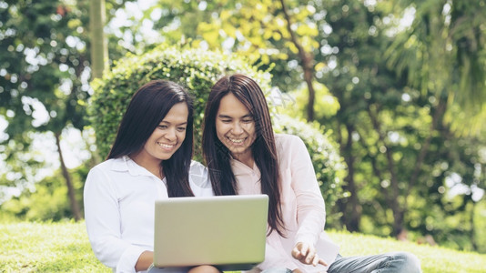 坐在公园绿色草地上谈恋爱的亚裔两名女使用笔记本电脑在公园绿色草原上聊天友谊女孩们聊天和看笔记本电脑规划如何做小型商业绿色花园享受背景图片