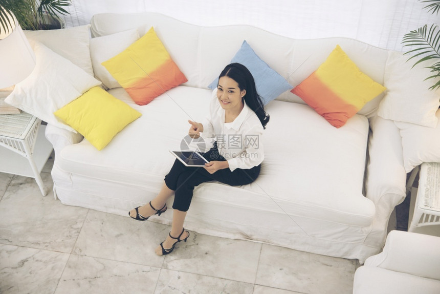人们工作室年轻美丽的亚洲女顶视图看着坐在白色沙发上的相机使用平板电脑智能手机浏览网站阅读文章和在线内容成人图片