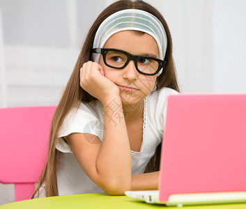 可爱的小女孩坐在桌子上带着粉红色笔记本电脑和戴眼镜坐着黑发美丽的图片