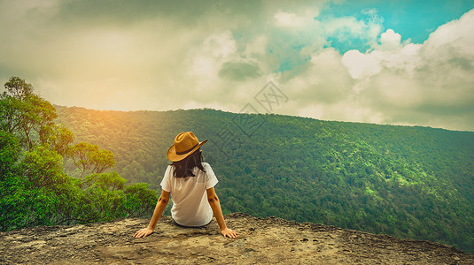 坐在悬崖假期户外身戴帽子坐在山顶悬崖的年轻旅行女青心情放松观看美丽的森林景色蓝天空和云彩风景在度假亚洲妇女独自旅行时士背景