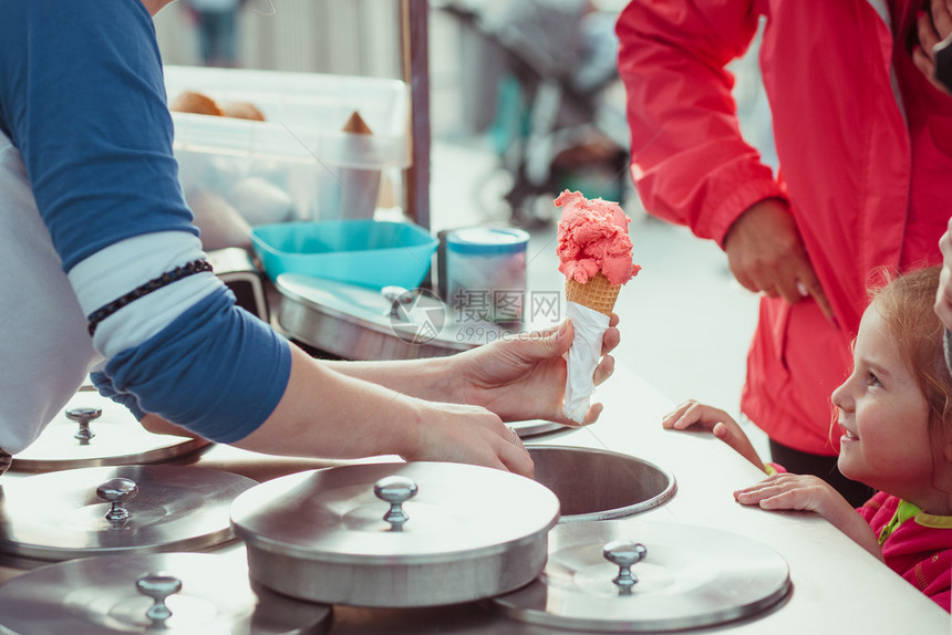 享受粉色的夏天小女孩等着冰淇淋妈在街边的糖果店里给女儿买冰淇淋年轻女孩把一勺粉红色冰淇淋送给一个锥体图片