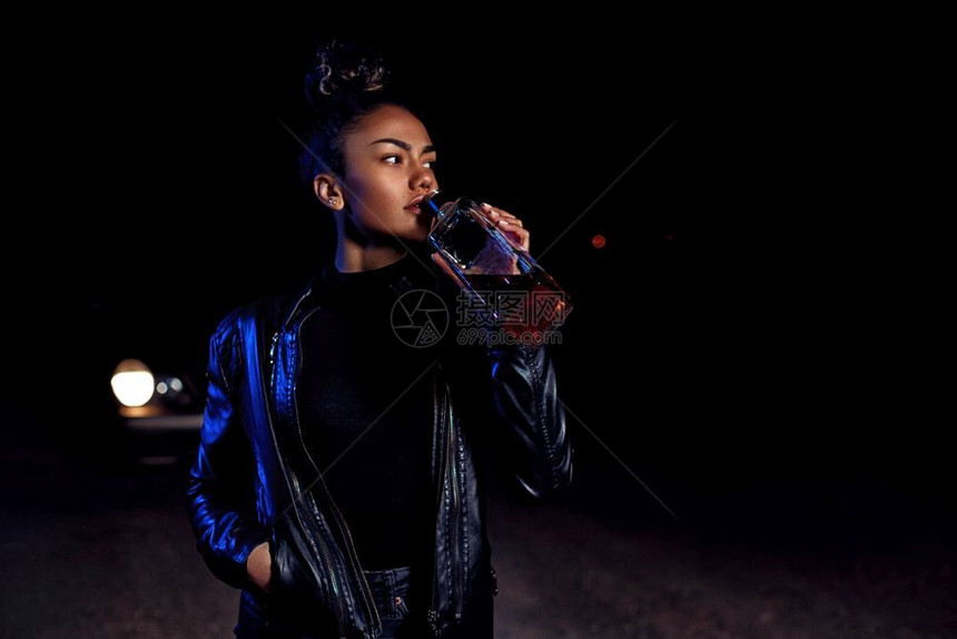 身穿皮夹克和黑衣服的非洲醉酒年轻黑人穆拉托女孩在一条废弃沙路上在汽车灯光和街道照亮下夜里喝着一瓶威士忌女酒精一种图片
