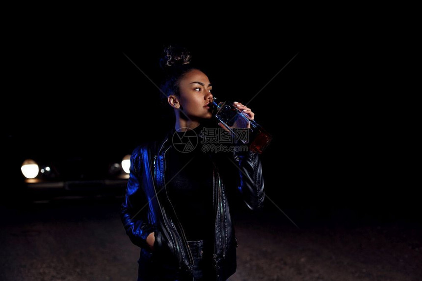 年轻的身穿皮夹克和黑衣服的非洲醉酒年轻黑人穆拉托女孩在一条废弃沙路上在汽车灯光和街道照亮下夜里喝着一瓶威士忌非洲人戏剧图片