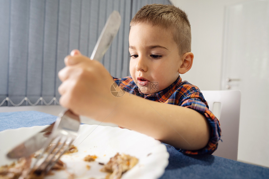 坐在桌边吃饭午餐或晚的幼小孩子肖像他们用叉子举着当家里的面朝前看着刀切板子食物男生晚餐图片