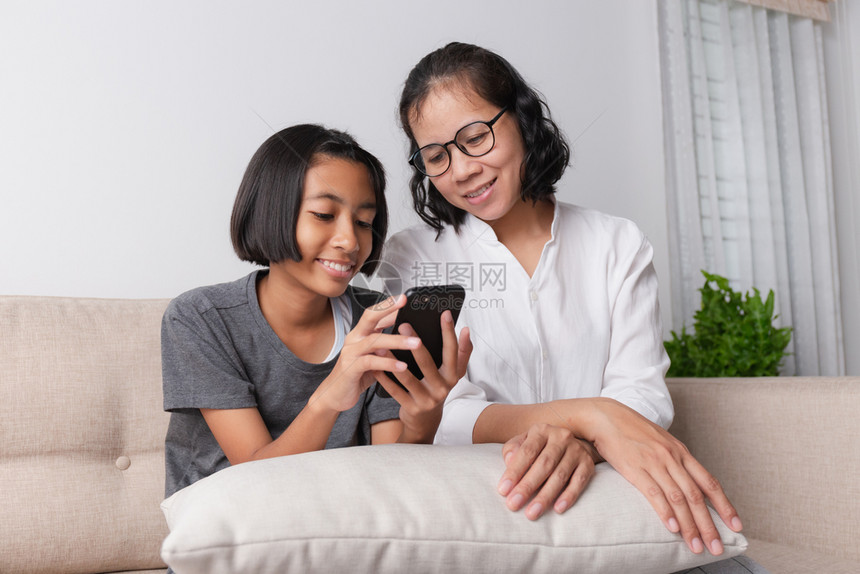 亚裔母亲和女儿在家中沙发上坐着时使用智能手机微笑与淑女抓住触碰图片