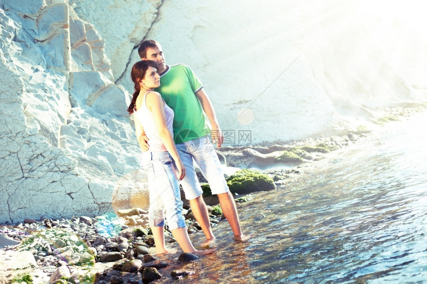 耀斑反射户外在岩石海岸的年轻夫妇站在脚踝深的水中看着远处图片