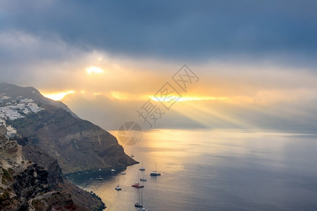 希腊桑托里尼太阳射线卡路德拉上空的重云照亮了海湾和数艘停靠的游艇圣托里尼卡尔德拉和穿透重云的太阳射线日出悬崖景观背景图片