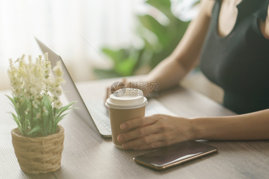 年轻妇女用一次咖啡杯喝并在工作桌上使用电脑笔记本办公室工作场所女图片