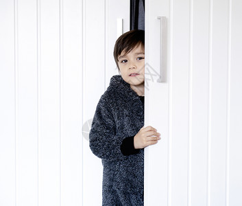 幸福杰米乐趣穿着毛睡衣的可爱孩子站在衣柜后面小男孩拿着白色门微笑地看着照相机儿童躲在衣橱里玩捉藏和躲在衣柜里图片