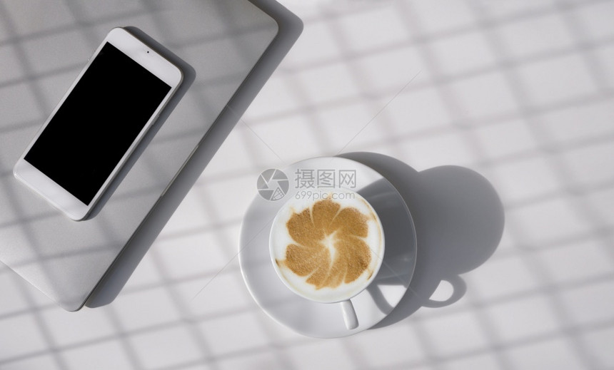 泡沫在咖啡店用空白智能手机和灰色笔记本电脑在桌子顶端的白陶瓷杯中热拿铁艺术咖啡表面的阳光和影子图案热的电话图片