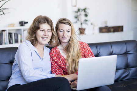 信用两名年轻妇女坐在起居室沙发上时使用计算机钱买方图片
