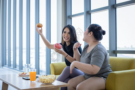 超重妇女和亚洲孩在家沙发上享受食物吃肥胖美味的不良图片