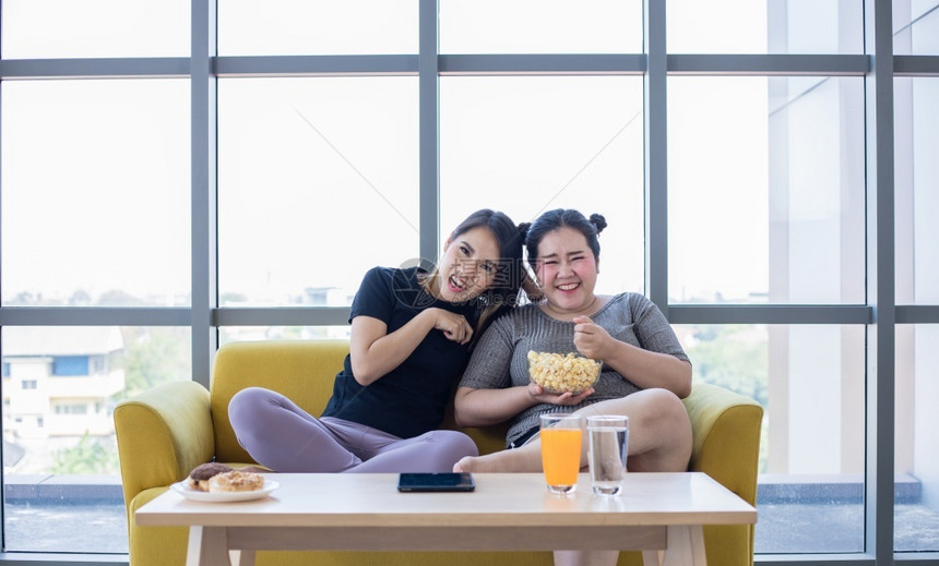 超重妇女和亚洲孩在家沙发上享受食物吃一顿饭暴食垃圾图片