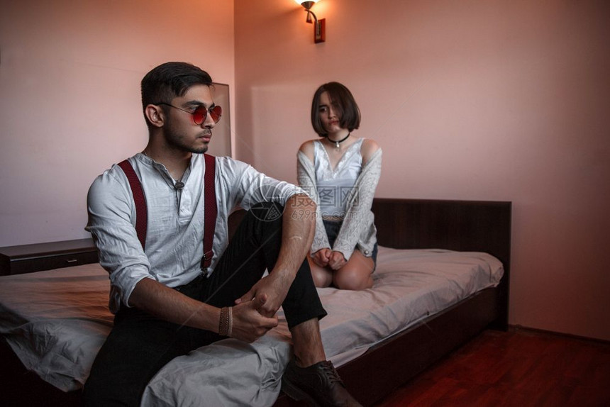 一个戴眼镜的年轻时尚男子和一个坐在床上的年轻女孩一个离女孩更近的男人有选择地关注她情绪化的重点失望图片