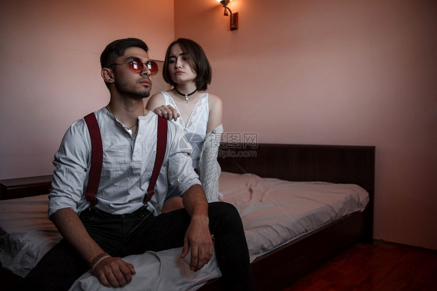 人们重点亲戚一个戴眼镜的年轻时尚男子和一个坐在床上的年轻女孩一个离女孩更近的男人有选择地关注她图片