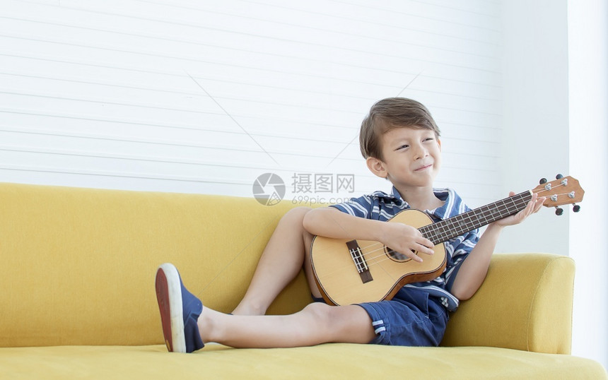音乐活的唱歌在家中客厅的黄色沙发上弹吉他时Caucasians男孩的肖像很快乐图片