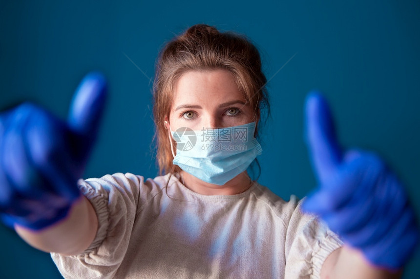 商业新冠一名年轻女子竖起大拇指戴着Covid19防护面罩和手套蓝色背景医学冠状一名年轻女子竖起大拇指蓝色背景冠状男人图片