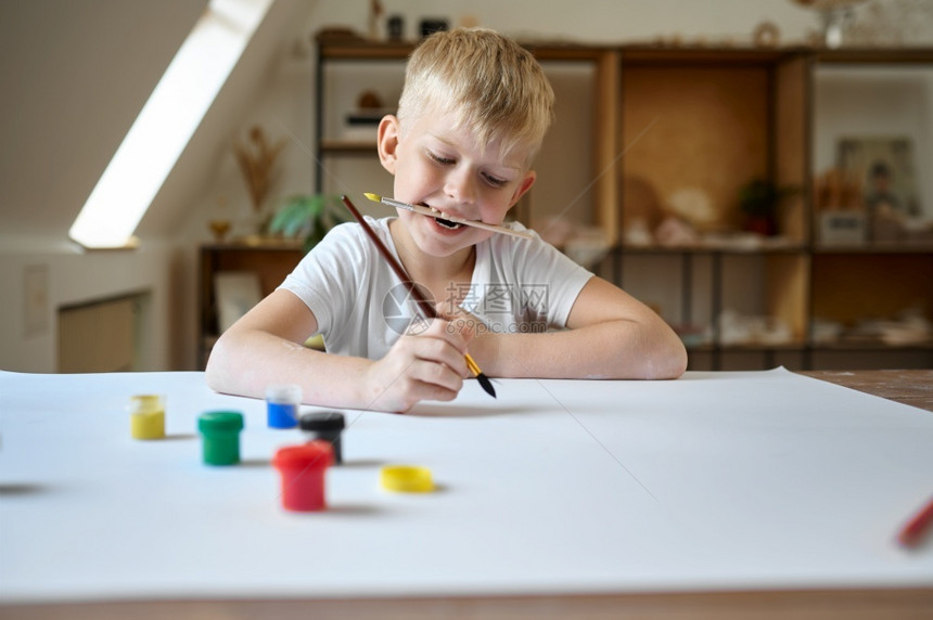 乐趣男坐在桌边的孩用古阿切画桌子在艺术学校习班上的孩子年轻画家愉快爱好乐童年与古阿希画桌子在讲习班上的孩子令人愉快图片