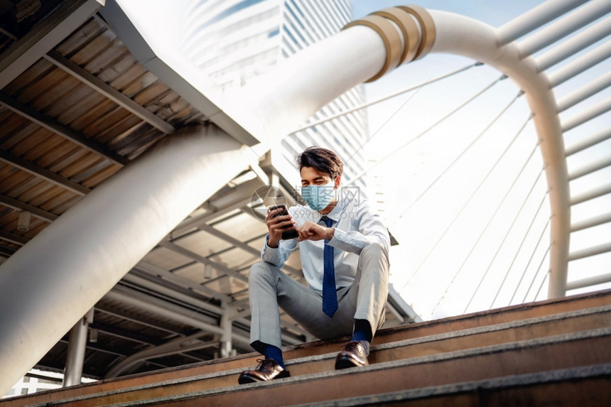 男人年轻亚洲商在市里穿着外皮面具和使用智能电话在新正常生活方式概念中坐在楼梯保健层的婴儿护理上生病的卫保健图片