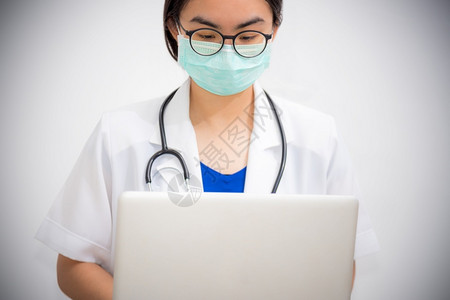看资料女医生灰色的亚洲年轻女医生佩戴眼镜和面具保护科罗纳的安全看笔记本电脑有听诊器的医生正在用一台电脑来阻止Covid19的流行医生正在用一设计图片