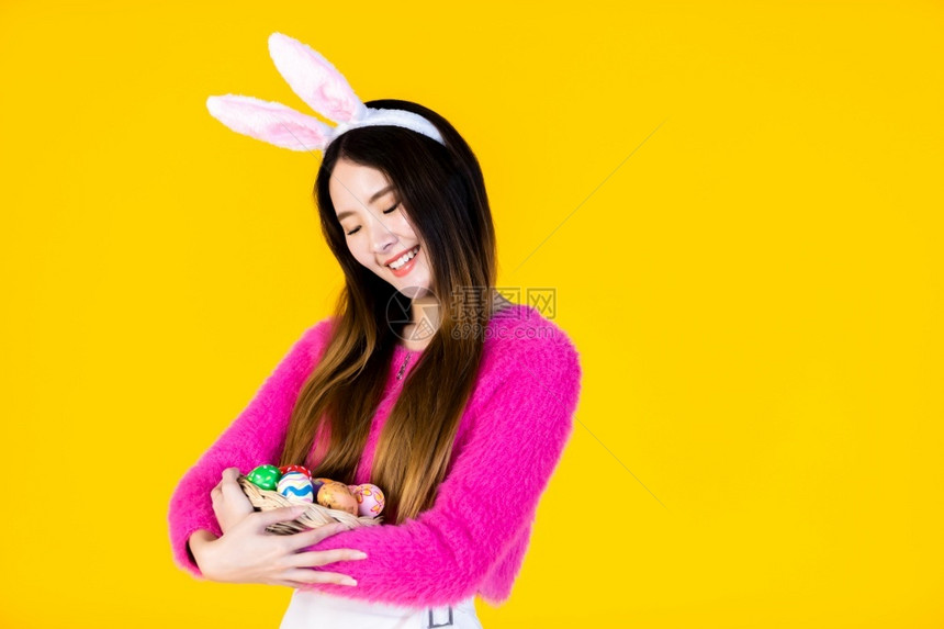 穿着兔子耳朵的亚洲快乐女青年手拿着一个篮子配彩色复活节鸡蛋在黄色空版间工作室背景上孤立无闻随意的夏天美丽图片
