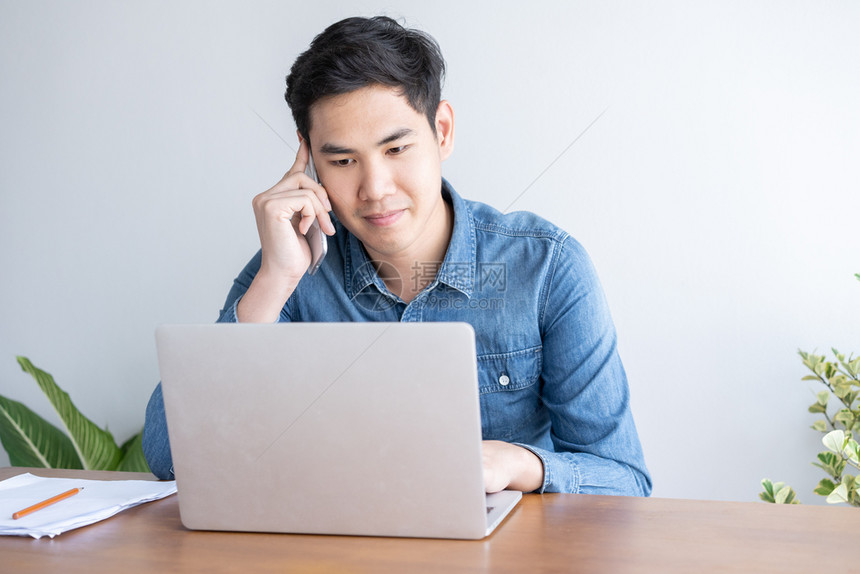 沟通年轻的青亚洲商人穿着蓝衬衫在手机上说话并在办公室用笔记本电脑工作话蓝色的图片