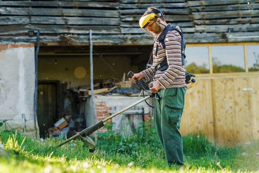 在职的年轻天主教男子农民园丁站在田野里手持连弦三角式汽油冲洗机切草在阳光明媚的一天在田里耕种村边风景的房子旁工作割草机械图片