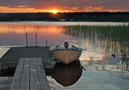 被搁浅小船木制的斯堪纳维亚树木在和平的多彩日落时由木制小船停靠在日落时被木制小船停靠在码头瑞典瓦尔姆兰一个湖上以宁静的水中天空反射着背景