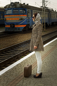 城市的延迟带眼镜和大衣的布鲁内特天主教妇女拿着手提箱在火车站等候运输图片