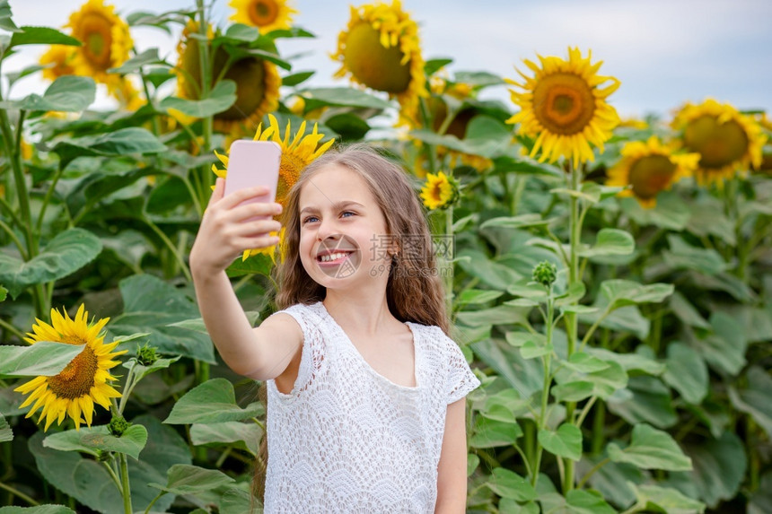 孩子飞女在智能手机上自拍脸带着微笑背景是一片向日葵花图片