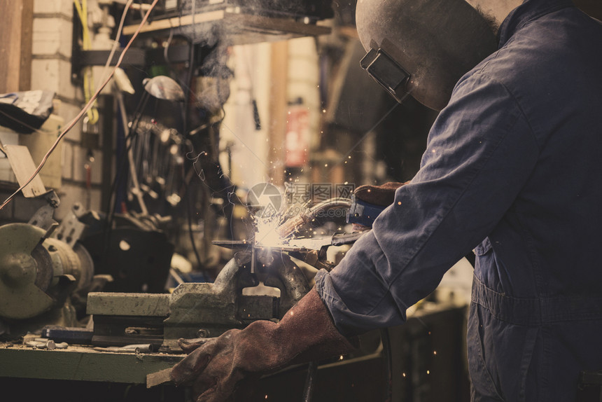 焊工正在用防护面罩焊接车库中的金属零件工业钢焊作机正在用防护面罩焊接车库中的金属零件工业钢焊机手动的气体工人图片
