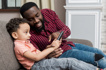 黑人父亲儿子使用智能手机沙发拥抱笔记本电脑枕头图片