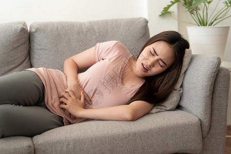 慢性浅表性胃炎说谎腹部月经疼痛的亚裔女和脸上表情达的亚裔女在睡家中沙发上时感到痛苦腹泻背景