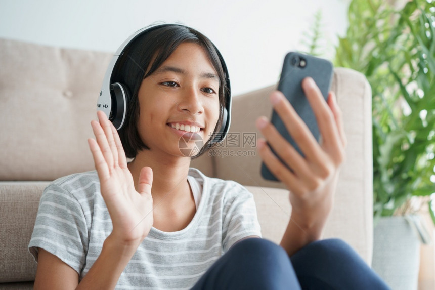 微笑亚洲小女孩视频电话在智能手机上戴耳头盔坐在家中起居室时手挥问候口令家庭放松和生活方式概念AC152L8青年细胞图片