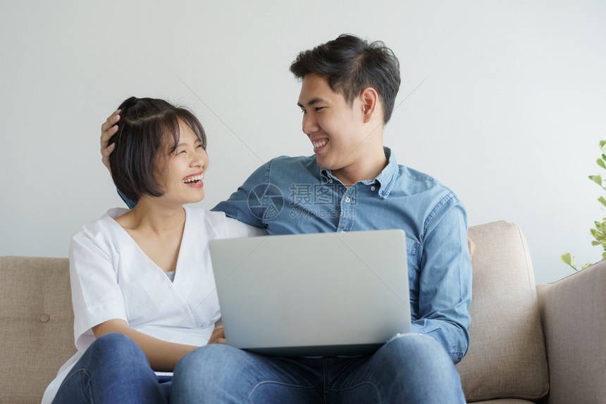 浪漫的人们情亚裔年轻夫妇在当代客厅的沙发上坐着取笑他们用笔记本电脑工作图片