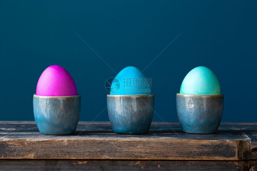 明亮的丰富多彩以蓝背景鸡蛋杯在木架子上涂满彩色闪亮复活节鸡蛋手用蓝背景的鸡蛋杯在木架子上涂满彩色鲜亮复活节鸡蛋假日弹簧卡复古设计图片