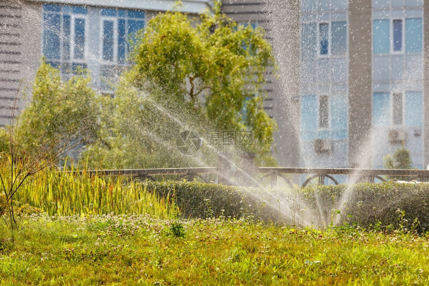 夏日炎热时花园灌溉洒水器在城市草坪上灌溉地而住宅楼的背景模糊场地冲洗户外图片