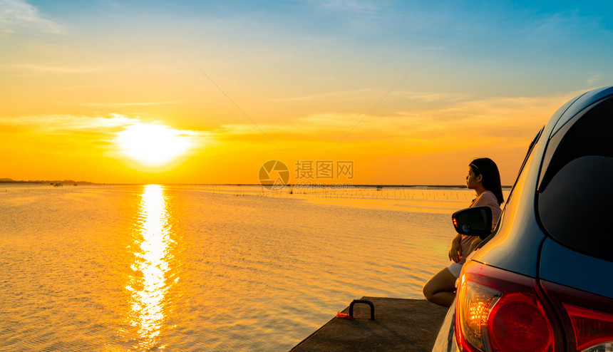 年轻亚洲女在日落时靠着蓝色紧凑的SUV运动车在海边倾斜头站上身穿T恤衫短裤和太阳镜友好水海滩图片