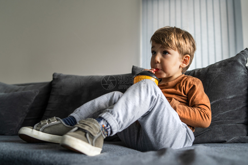 一种三四岁或小可爱的幼天主教男孩的肖像他们坐在床上或沙发在家里拿着一个塑料杯在家中独自一人喝着草汁或苏打水整天排靠边站着人们液体图片