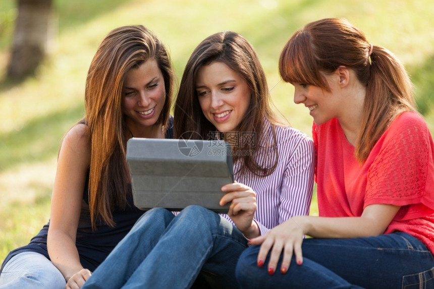 三个女孩坐在草地上愉快地看着平板图片