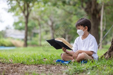 一名身戴卫生面具的白人男孩独自坐在一棵树下阅读本书电晕童年白种人图片