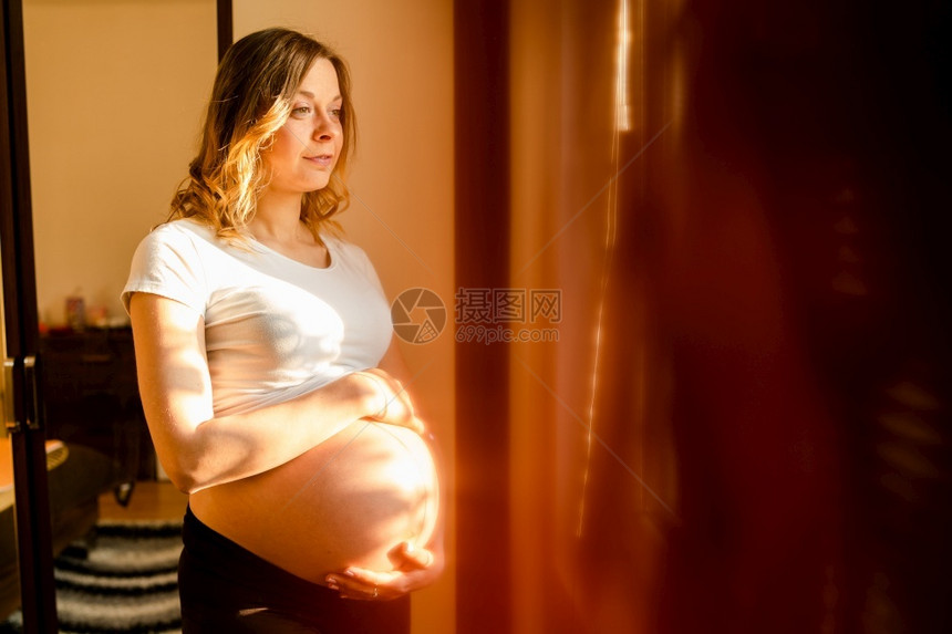 女士分娩妈年轻美丽的孕妇站在窗边看着她的肚子在等待婴儿的到来望着她肚子图片