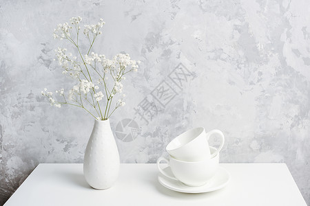 老的早晨结石花瓶中的一束小满天星和灰色混凝土墙上的桌子两个白色茶杯或咖啡复制空间前视图背景图片