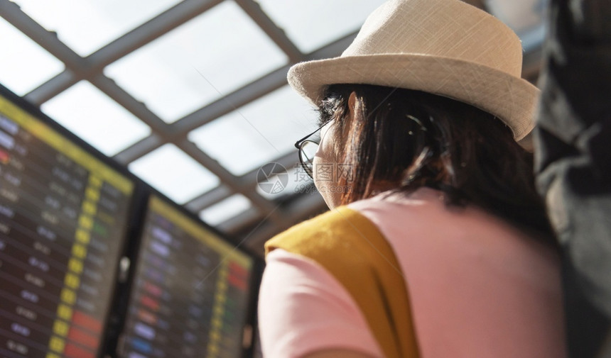 时间表背包客飞机场亚洲女游客戴帽子和穿粉红色衬衫在飞行日程安排上观察飞行时间后观女士旅客在机场起航候站等图片