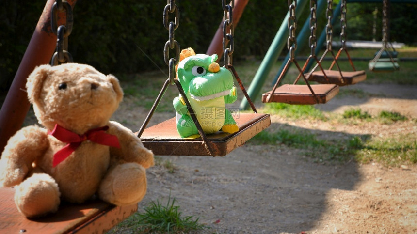 夏天玩具娃早晨有选择地关注可爱绿色的恐龙娃泰迪熊坐在公共园区游乐场秋千摆放图片