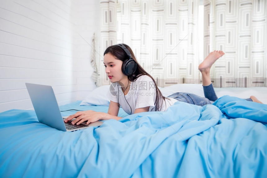 亚洲快乐的年轻妇女戴耳机听音乐歌曲观看网站在家床上用笔记本电脑在网上购物校对Soup穿女士移动的图片